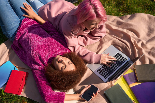 Вид сверху улыбающейся женщины со смартфоном, лежащей на траве рядом со своей спутницей на ноутбуке - Фото, изображение