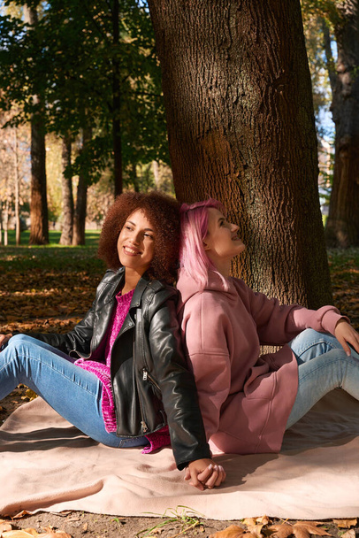 Femme souriante et sa compagne heureuse assise sur une couverture sous un arbre et tenant la main - Photo, image