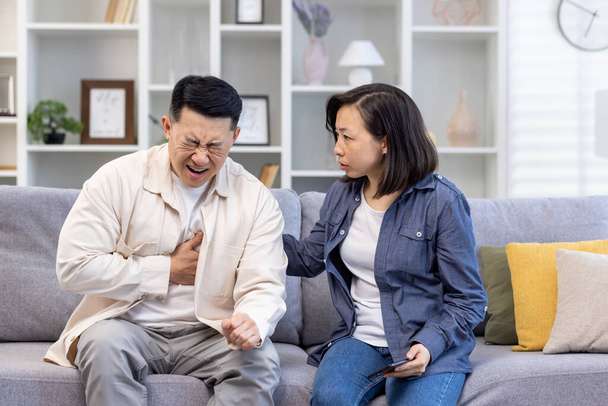 Young Asian family sitting on sofa at home. A man holds his hand to his chest, heart attack, stroke, panic attack, feels severe pain. Shocked woman tries to help, give first aid. - Valokuva, kuva
