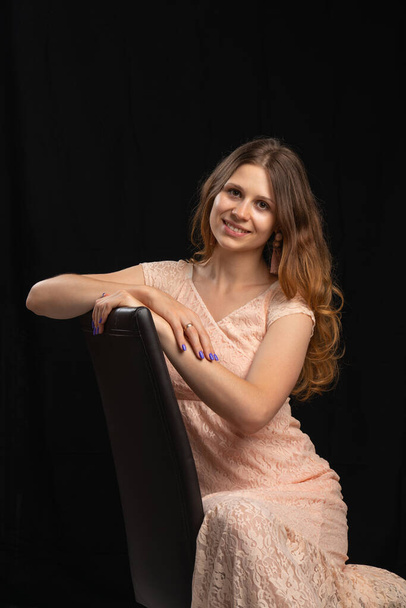 A young beautiful girl with dark long hair in a light pink dress sits on a black chair. black background.Studio shooting - Foto, Bild