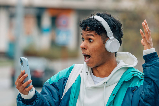 νεαρός άνδρας με ακουστικά και κινητό τηλέφωνο με έκπληκτη έκφραση - Φωτογραφία, εικόνα