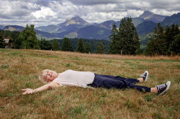 A mulher idosa deita-se entre as montanhas de braços estendidos, sobrecarregada de emoções. Bela paisagem. Envelhecimento ativo, paz interior e tranquilidade - Foto, Imagem