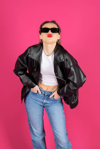 Cool biker girl wearing a leather jacket and jeans makes a kiss. Isolated on pink background. - Foto, Imagem