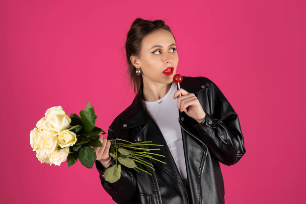 Δροσερό κορίτσι μοτοσυκλετίστρια με δερμάτινο μπουφάν και τζιν που ποζάρει με μπουκέτο λευκά τριαντάφυλλα και γλειφιτζούρι. Απομονωμένα σε ροζ φόντο. - Φωτογραφία, εικόνα