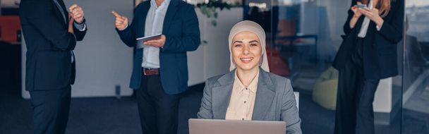 Smiling muslim businesswoman in hijab working on computer on colleague background - Φωτογραφία, εικόνα