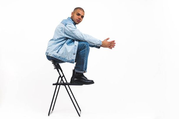 Young adult male model in denim set sitting on a chair in unconventional way. Dangerous poses during fashion photoshoot. High quality photo - 写真・画像