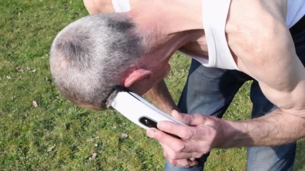 un hombre de mediana edad de pelo gris se afeita el cabello con un cortador en un jardín en un césped verde, imágenes de alta calidad 4k - Imágenes, Vídeo