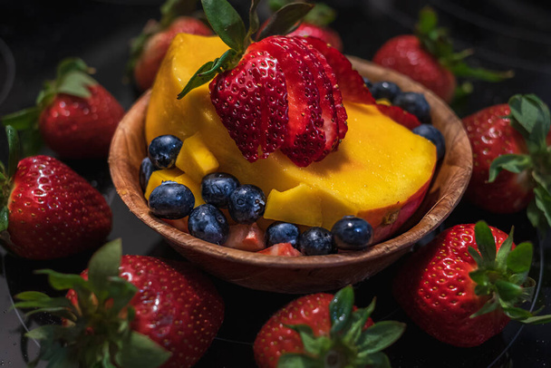 Wooden bowl with fresh fruits salad on dark background. Bowl of fruit salad with berries and mango. Nobody, selective focus, blurred - Photo, image