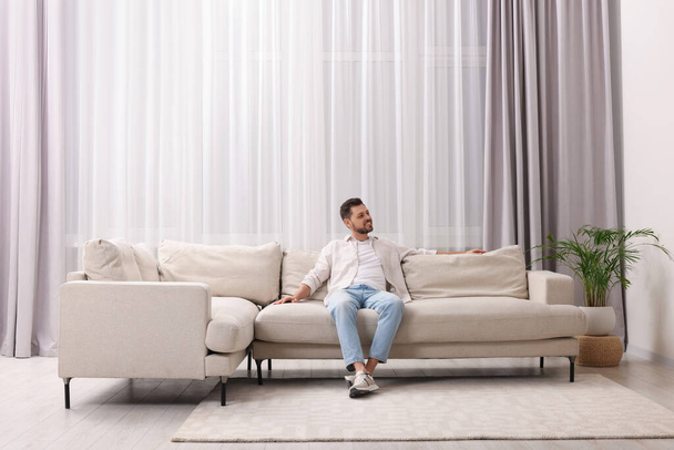 Happy man resting on sofa near window with beautiful curtains in living room - Foto, Bild