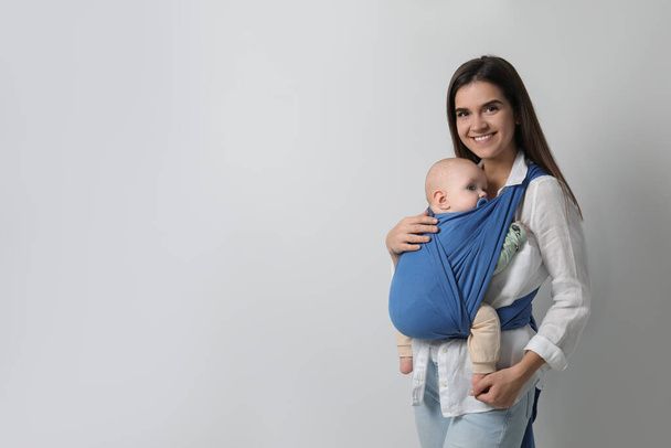 Mother holding her child in sling (baby carrier) on light grey background. Space for text - 写真・画像