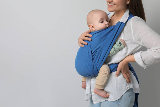 Mother holding her child in sling (baby carrier) on light grey background, closeup. Space for text - Fotó, kép