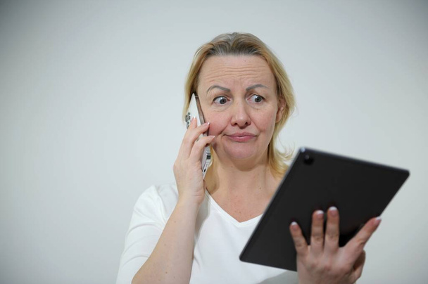 Stressful female entrepreneur communicates via cell phone during work at digital tablet, displeased hear bad news, blows cheeks in dissatisfaction, doesnt know how make task sits at desktop - Foto, afbeelding