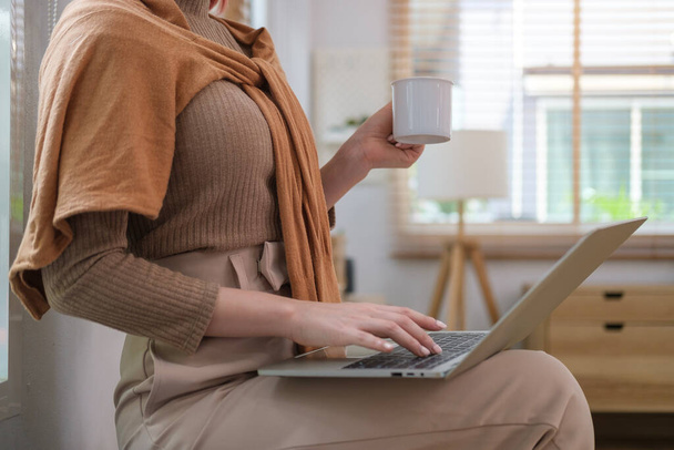 Cropped shot of modern young woman in stylish clothes holding coffee cup and using laptop at home. - Фото, изображение