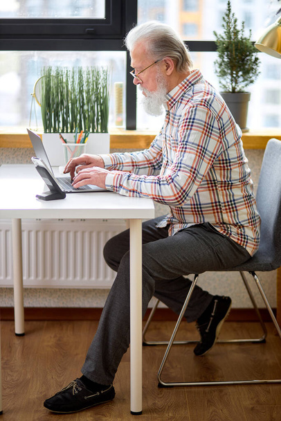grandfather checking social media and reading news or shopping online while sitting at home. confident senior gray-haired male in casual shirt sit in cozy bright room websurfing, working, studying - Fotografie, Obrázek