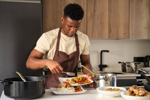 Junger schwarzer Mann serviert Kochbananen auf Tomatensauce gebratenes Hühnerhack und Reisteller in einer Küche. - Foto, Bild