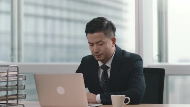 uomo d'affari asiatico seduto alla scrivania in ufficio moderno guardando computer portatile che celebra il successo e il raggiungimento - Filmati, video