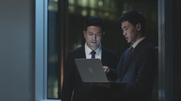 due uomini d'affari asiatici in piedi vicino alla finestra in ufficio di notte avendo una discussione utilizzando il computer portatile - Filmati, video