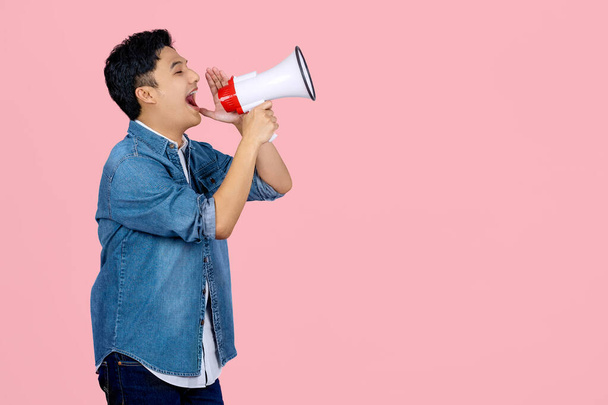 Handsome Asian man in blue shirt shouting in megaphone announcing a sale. Hurry up isolated on pink background studio photo - Photo, Image