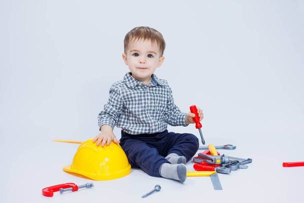 Un niño pequeño con una camisa con un casco y una herramienta de juguete. Fondo blanco. Herramienta de construcción de juguetes para niños - Foto, imagen