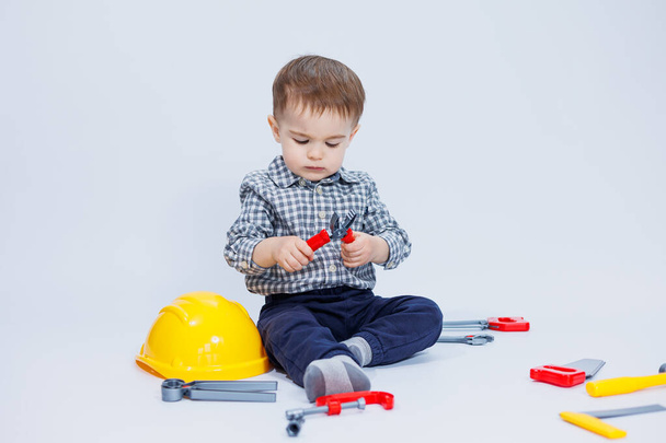 Un chico lindo con una camisa en la imagen de un constructor, sobre un fondo blanco. Infancia, aprendizaje, trabajo, juegos, carrera, imitación. Ayudante del pequeño papá. - Foto, Imagen