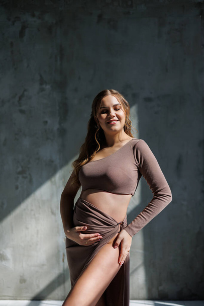 Μια ευτυχισμένη έγκυος γυναίκα με καφέ κοστούμι ποζάρει και χαϊδεύει την κοιλιά της. Έγκυος έγκυος γυναίκα. Ευτυχισμένη εγκυμοσύνη - Φωτογραφία, εικόνα