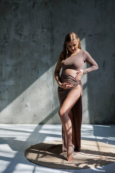 Счастливая молодая беременная женщина в коричневом нежном костюме позирует и гладит живот. Беременная женщина. Счастливая беременность - Фото, изображение