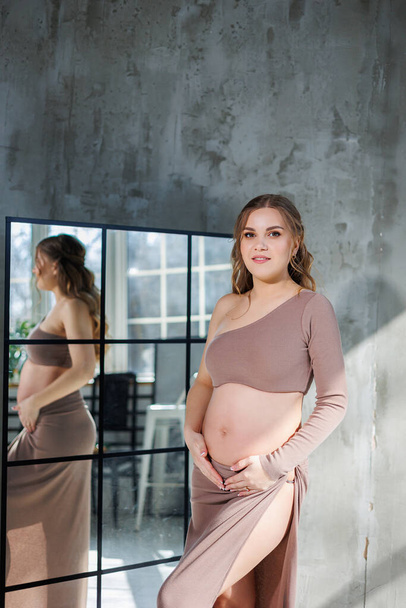 A pregnant woman in a skirt hugs her stomach with her hands. Fashion pregnant woman. Full length portrait on gray background. Happy pregnancy - Фото, изображение