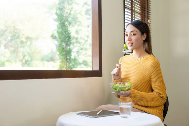Młoda piękna Azjatka szczęśliwie jedząc sałatkę warzywną w domu na wakacjach, jedząc koncepcję wellness. - Zdjęcie, obraz
