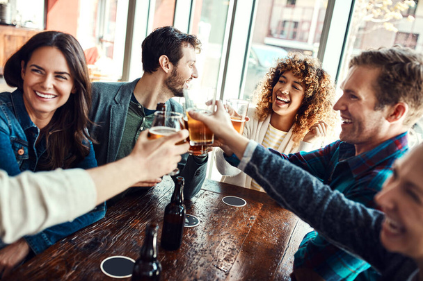 Bier is wat ons samenbrengt. een groep vrienden die een toost uitbrengen terwijl ze zich vermaken in een bar - Foto, afbeelding