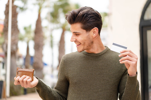 Joven hombre caucásico al aire libre sosteniendo billetera y tarjeta de crédito con expresión feliz - Foto, imagen