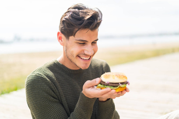 Young caucasian man at outdoors holding a burger - Φωτογραφία, εικόνα