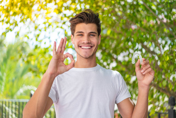 Young caucasian man holding invisible braces at outdoors showing ok sign with fingers - Fotoğraf, Görsel