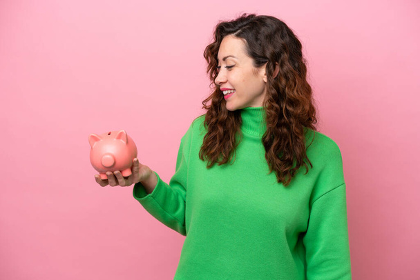 Young caucasian woman holding piggybank isolated on pink background with happy expression - Foto, Imagen