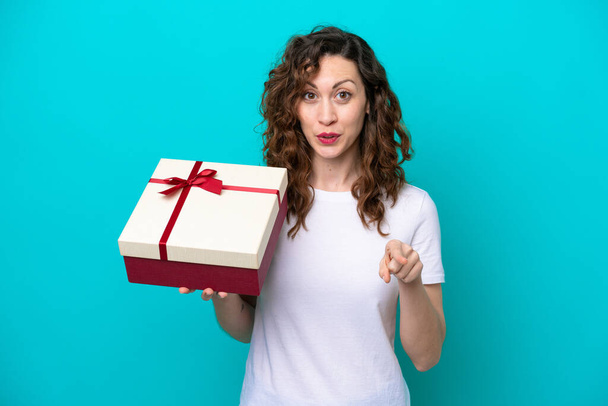 Young caucasian woman holding a gift isolated on blue background surprised and pointing front - 写真・画像