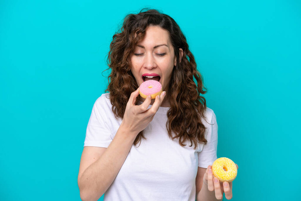 Young caucasian woman isolated on blue background eating a donut - Photo, Image