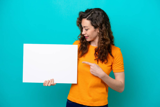 Молода біла жінка ізольована на синьому фоні, тримаючи порожній плакат з щасливим виразом і вказуючи на нього
 - Фото, зображення