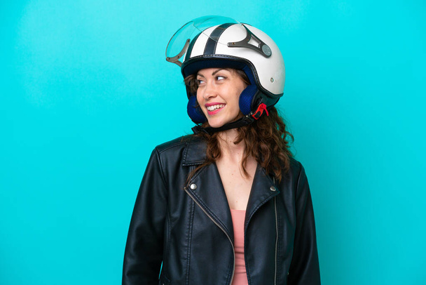 jonge Kaukasische vrouw met een motorfiets helm geïsoleerd op blauwe achtergrond kijken naar de zijkant en glimlachen - Foto, afbeelding