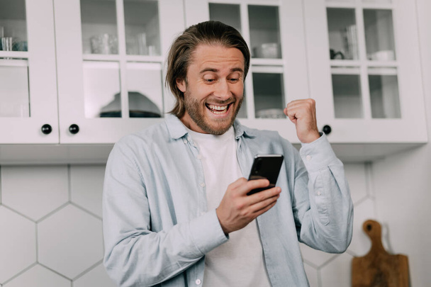 A young man rejoices in success looking at his smartphone at home. Lucky bet, stock market, crypto market. - Fotografie, Obrázek