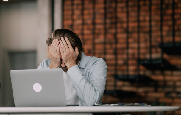 Stressful office job finally turns young man into a burnout. Stressed, tired, exhausted male worker overwhelmed with problems sitting at working desk with laptop computer and holding hand on his face - Foto, afbeelding