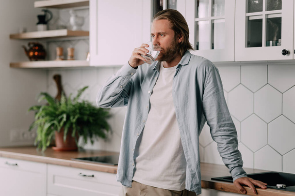 attractive young man stand drinking water . Handsome male drink or take of mineral natural in glass after wake up in morning for health and wellbeing in kitchen in house - Valokuva, kuva