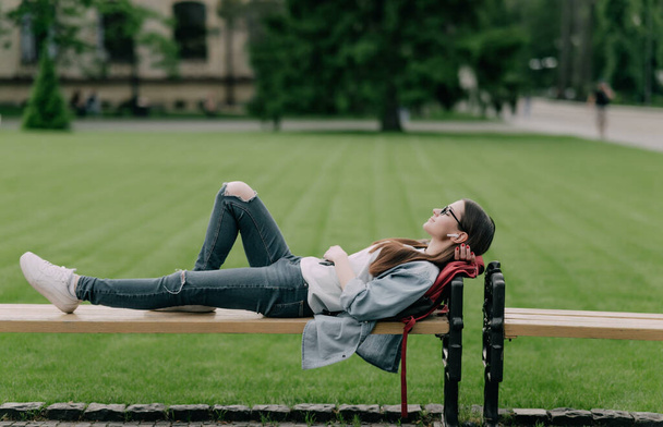 Young beautiful natural looking student lying on a banch in a campus listening to music - Foto, imagen