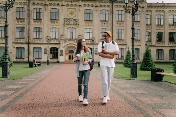 Two international stylish students walk near the campus and smile. Friendly African American guy and Caucasian girl walk together near the university. - Foto, Imagem