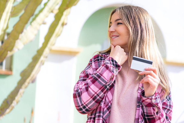 Joven mujer rumana bonita sosteniendo una tarjeta de crédito al aire libre pensando en una idea y mirando al lado - Foto, imagen