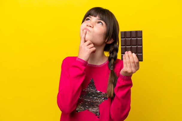 Μικρή καυκάσια κοπέλα με σοκολάτα απομονωμένη σε κίτρινο φόντο που έχει αμφιβολίες ενώ κοιτάζει ψηλά - Φωτογραφία, εικόνα