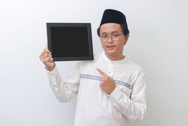 Retrato de un joven musulmán asiático sosteniendo una pizarra con la mano en punta. Espacio en blanco para tipografía o texto a rellenar. Imagen aislada sobre fondo blanco - Foto, imagen