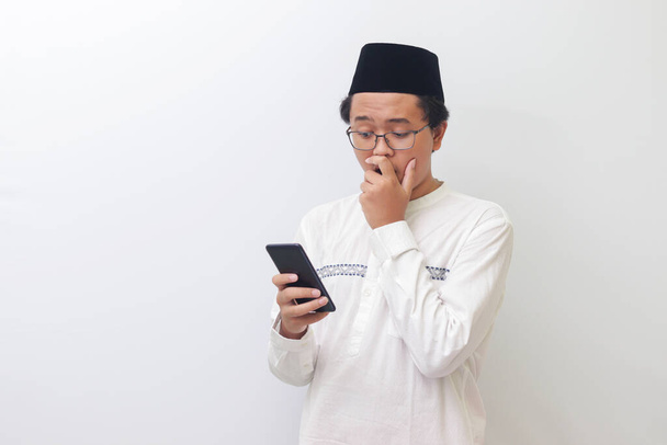 Retrato del joven musulmán asiático sosteniendo el teléfono móvil y cubriendo la boca con expresión conmocionada en la cara. Imagen aislada sobre fondo blanco - Foto, imagen