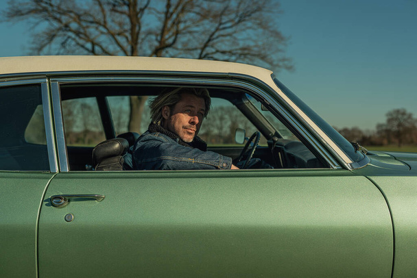 Homem com cabelo loiro em jeans jaqueta senta-se no lado do passageiro de um carro muscular americano vintage olhando pela janela. - Foto, Imagem