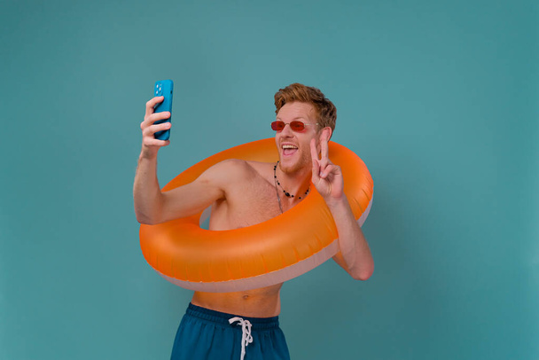 Cheerful shirtless man standing isolated over blue background, taking a selfie with inflatable ring. Summer vacation concept - Fotó, kép