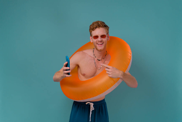 Rudowłosy mężczyzna w okularach przeciwsłonecznych stojący z pomarańczowym gumowym pierścieniem i wskazujący na smartfona. Koncepcja letnich wakacji na niebieskim tle. - Zdjęcie, obraz