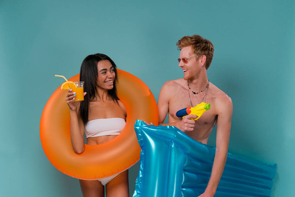 Pool party fun. Multiethnic Couple with inflatable rings having fun shooting with water guns, blue studio background. Summer vacation sea sun tan concept - Foto, Bild